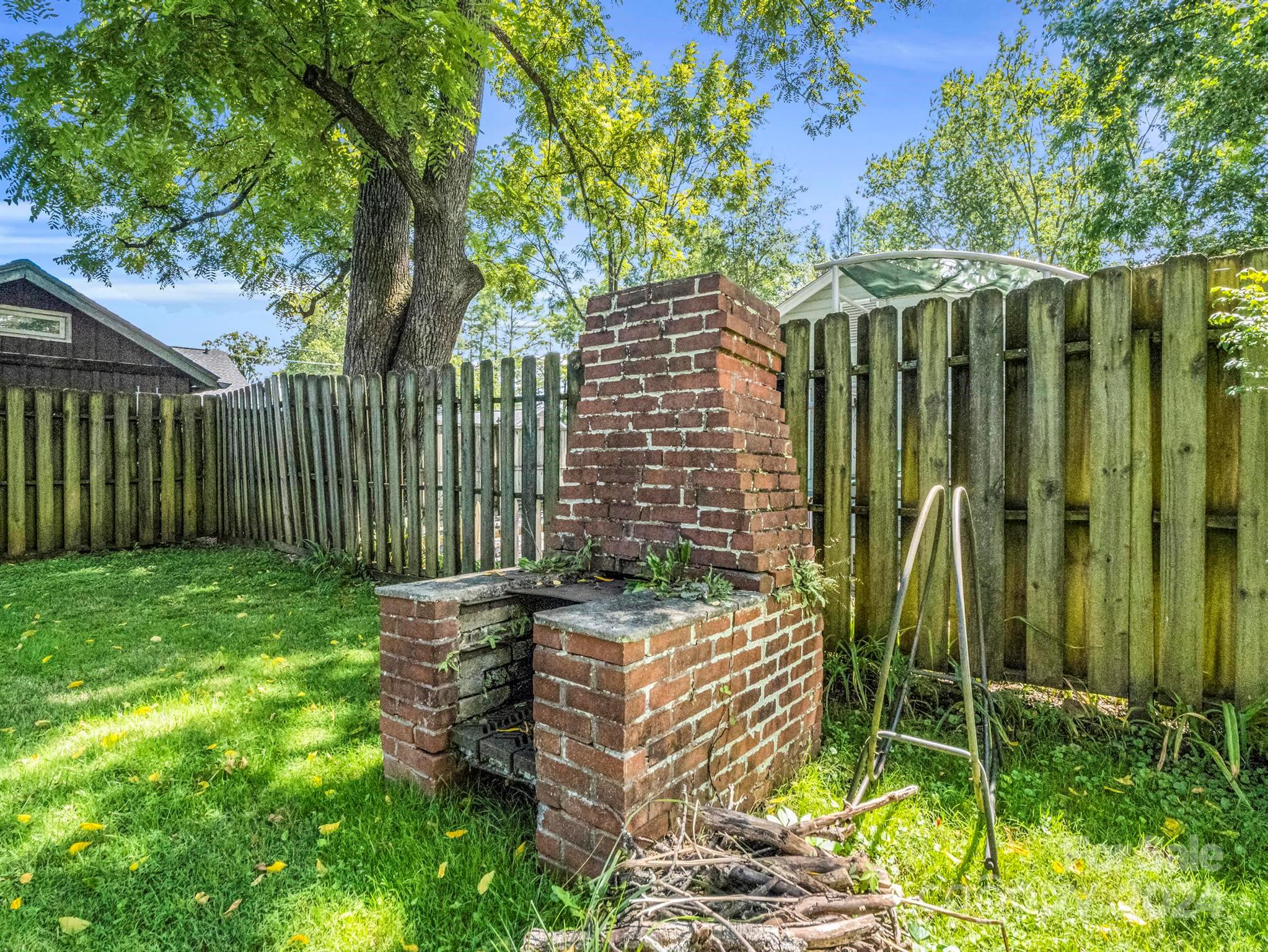 Backyard with a brick outdoor fireplace, wooden privacy fence, mature trees, and grassy lawn.