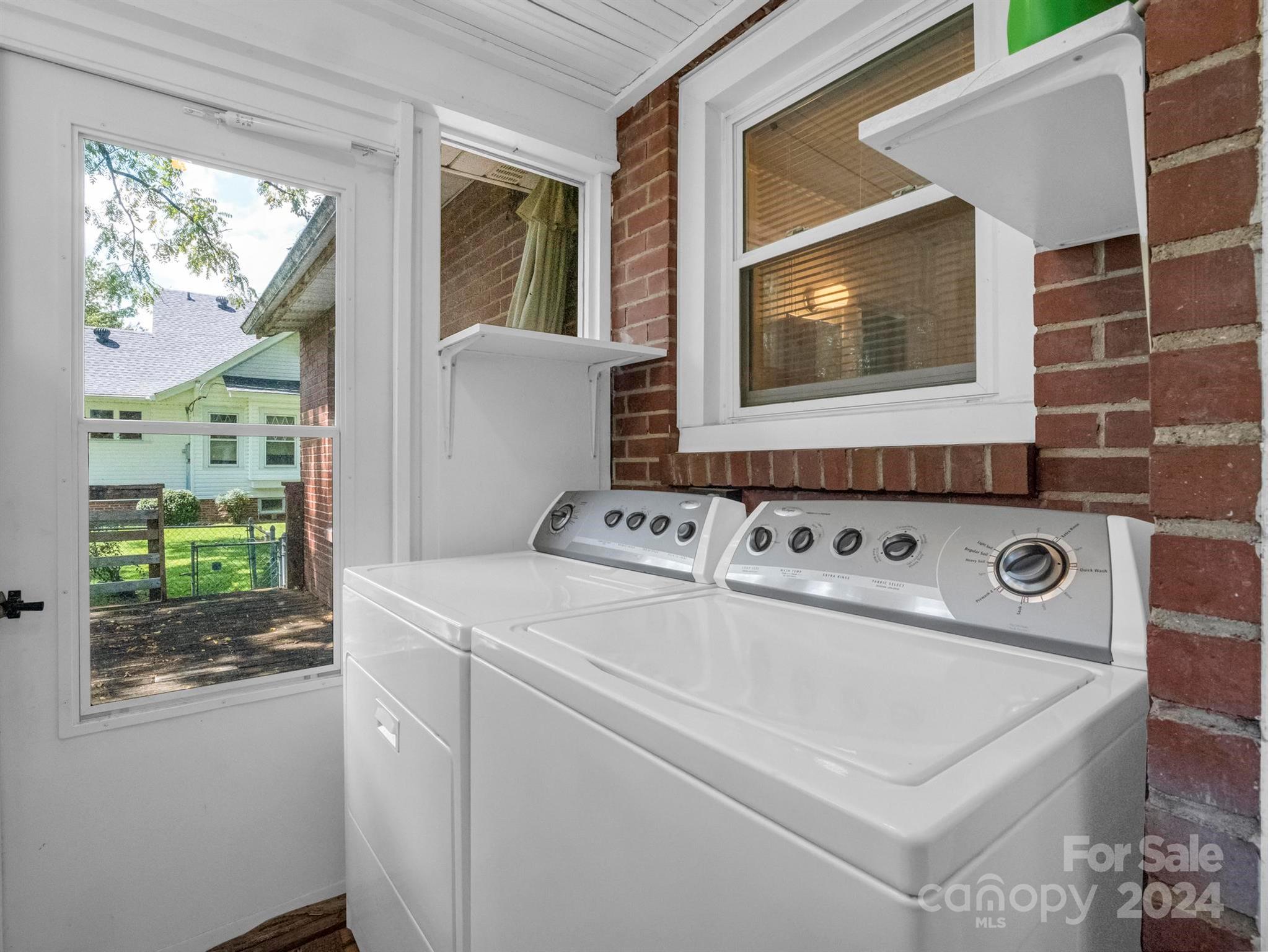 Laundry room with a white washer and dryer beside exposed brick walls and large windows.