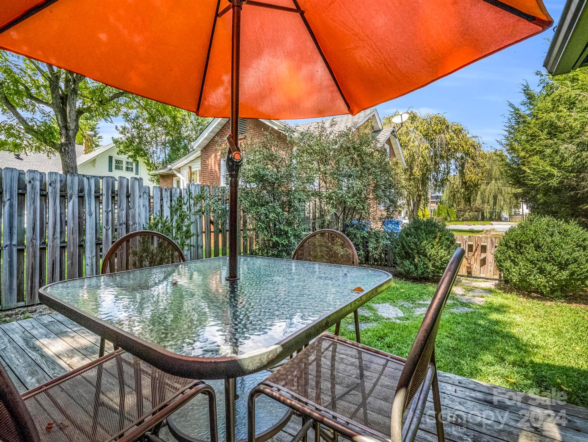 Backyard patio with a glass-top dining table beneath a large orange umbrella, beside a fenced lawn.