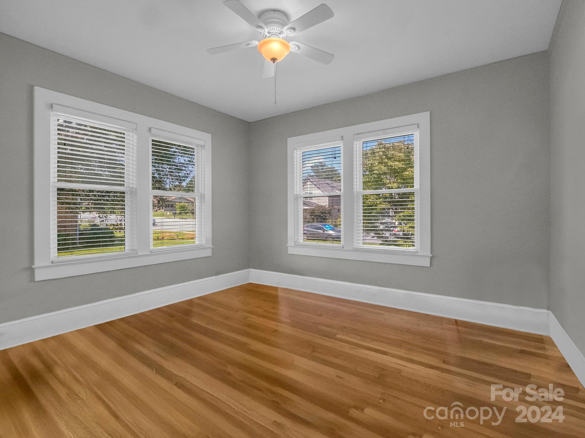 Empty room with polished hardwood floors, gray walls, and four windows with white blinds.