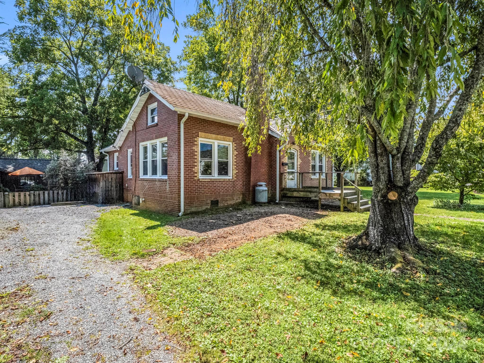 Red-brick house with a raised wooden deck, gravel driveway, and large leafy tree in the yard.