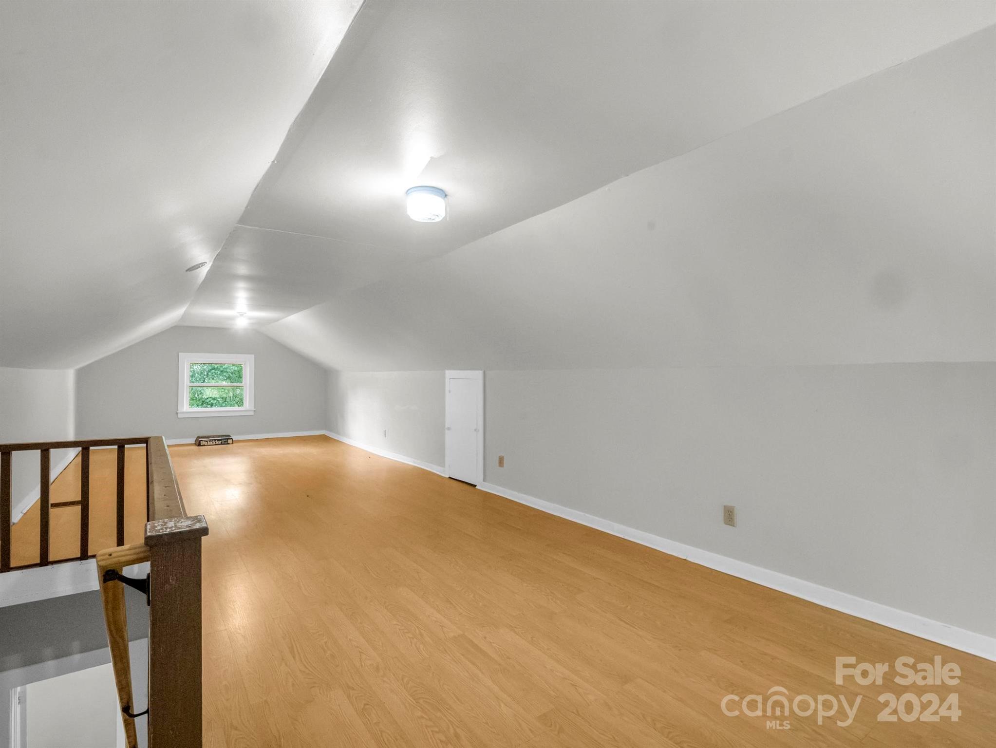 Vacant attic loft with sloped ceiling, wood flooring, small window, and stair railing.
