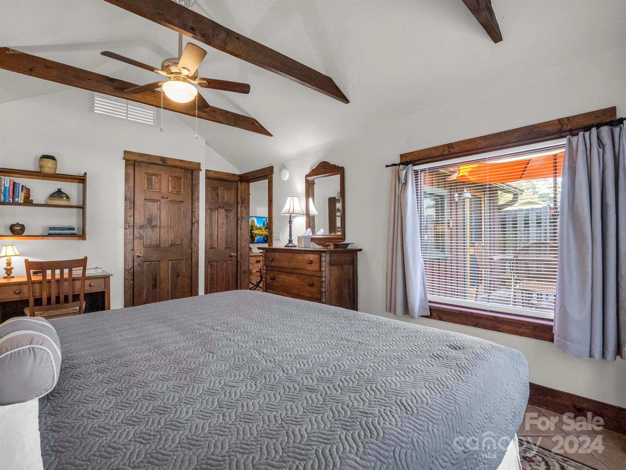 Bedroom with a gray-covered bed, exposed wood ceiling beams, and a large curtained window.