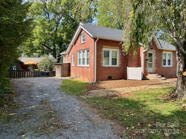 Red-brick house with white trim beside a gravel driveway, mature trees, and a fenced yard.