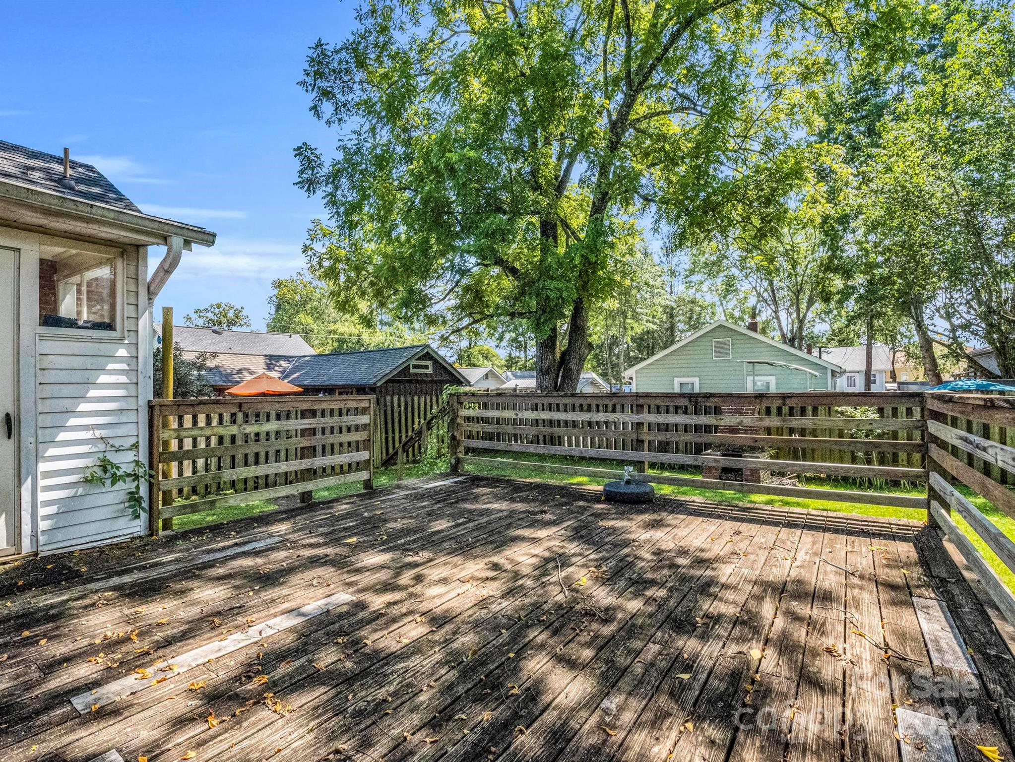Weathered wooden deck with a railing beneath a large leafy tree, with neighboring houses beyond.