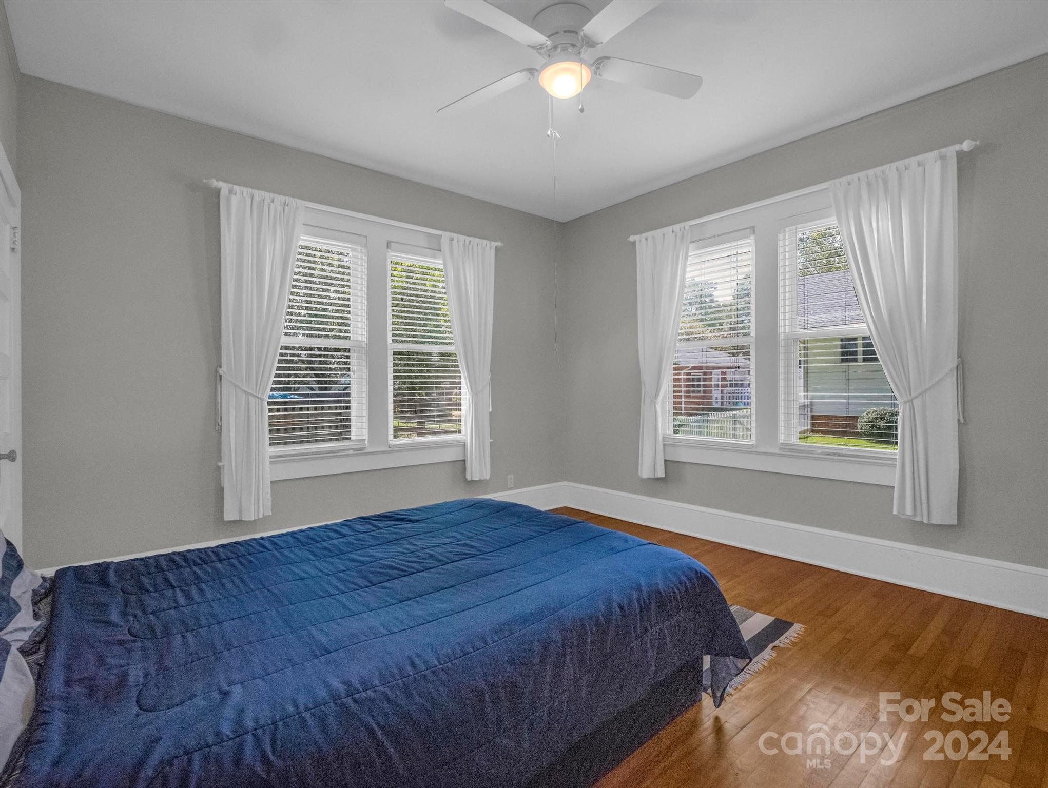 Bedroom with a blue-quilted bed, hardwood floors, and two window sets with white curtains.