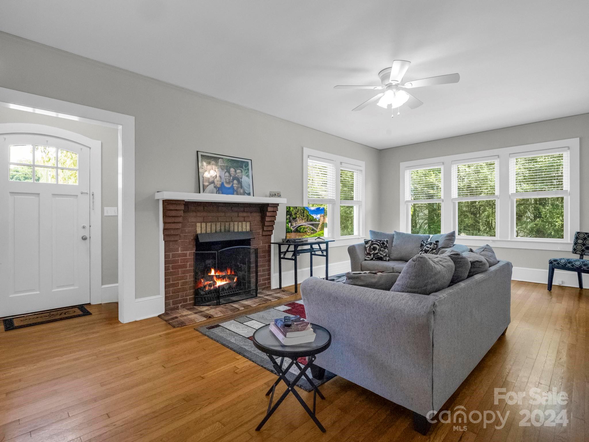 Living room with a brick fireplace, gray sectional sofa, hardwood floors, and large windows.