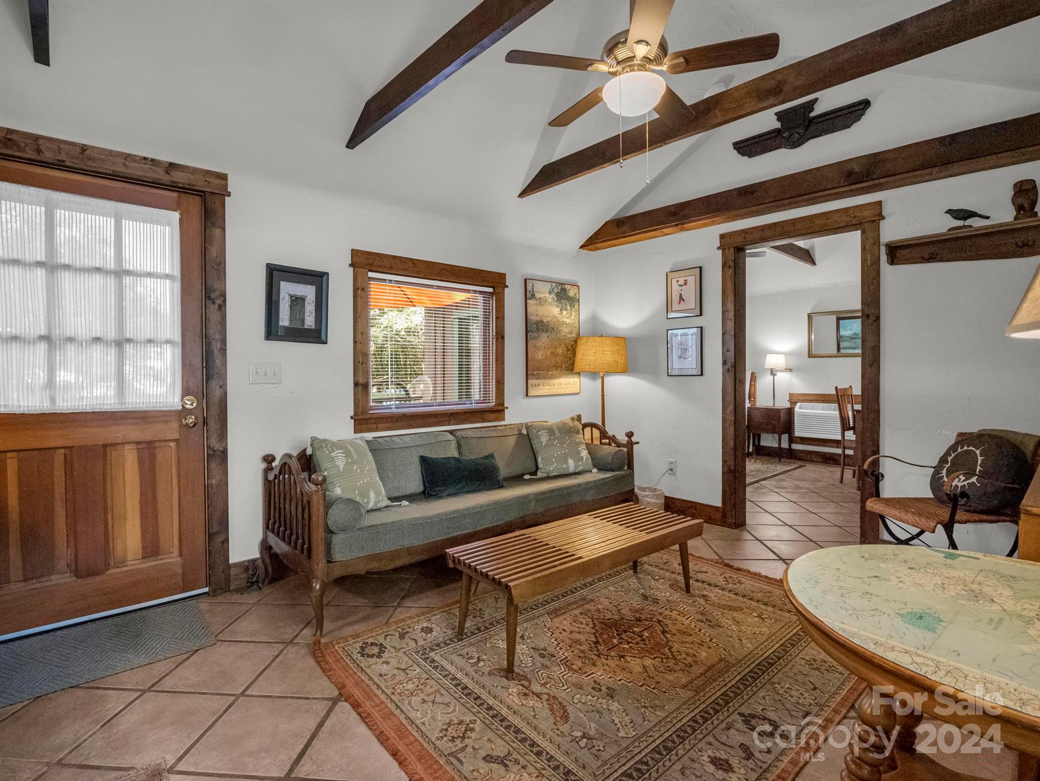 Living room with exposed wood beams, a gray sofa, tiled floor, and doorway to a dining area.