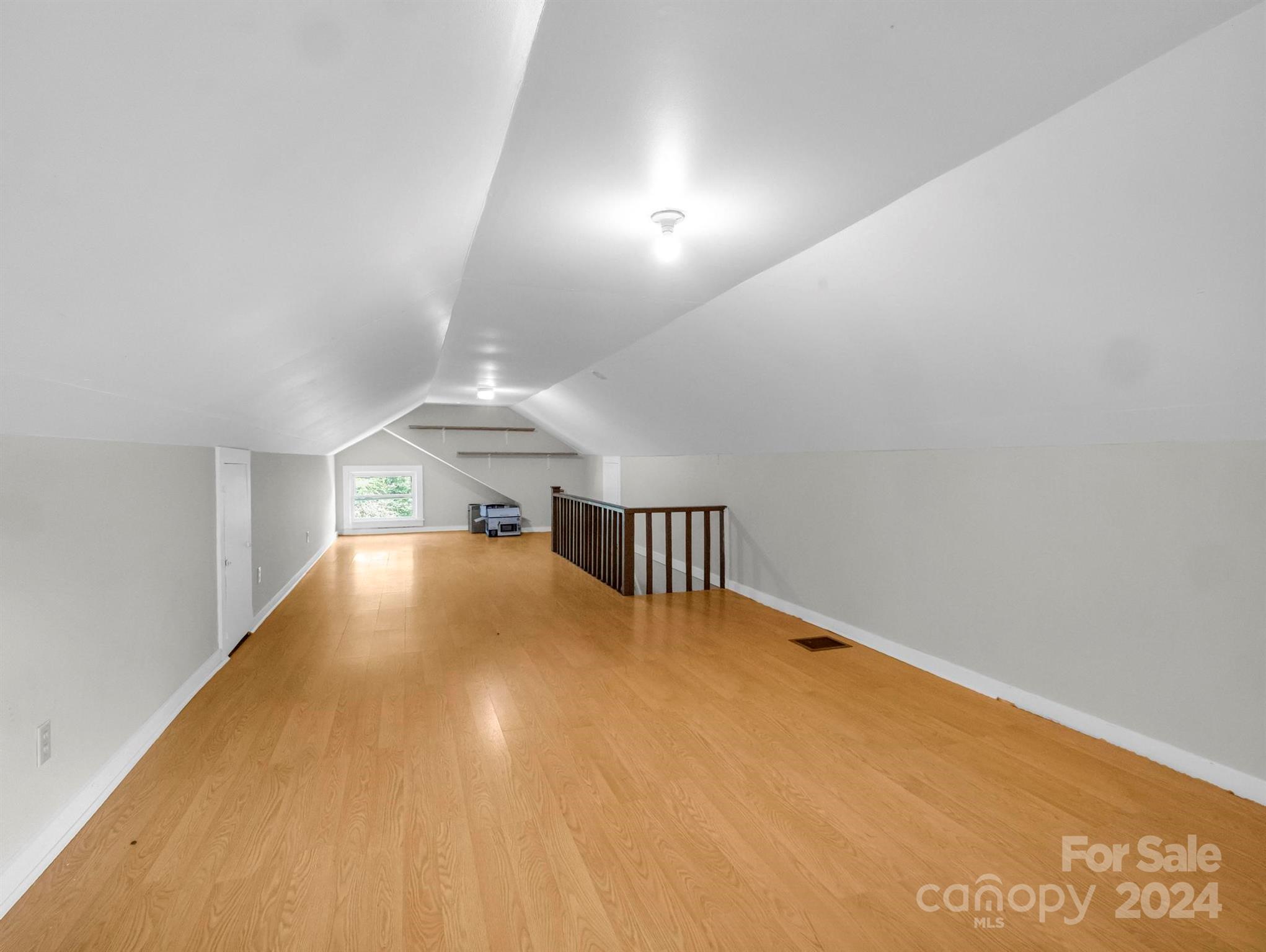 Empty upper-level room with a sloped ceiling, light wood floors, and a wooden stair railing.
