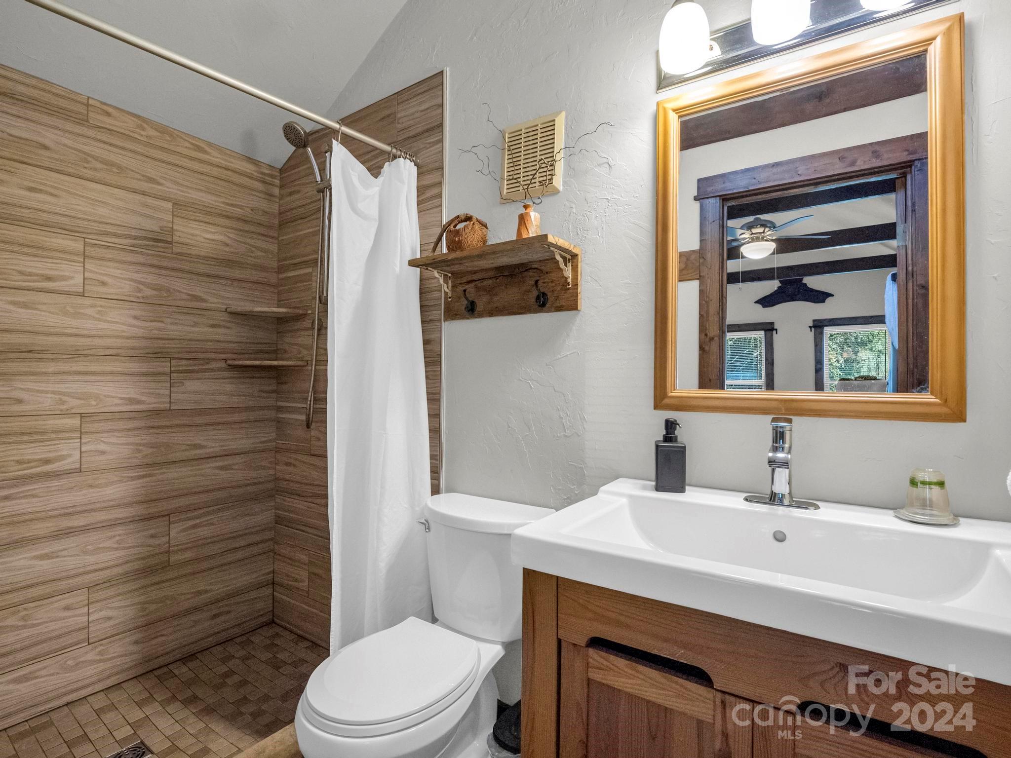 Bathroom with wood-look tiled shower, white curtain, toilet, and wood vanity beneath a framed mirror.