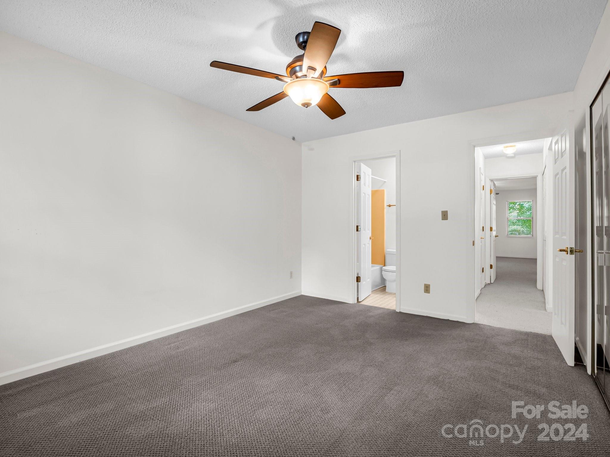 Empty carpeted bedroom with a ceiling fan, mirrored closet doors, and open bathroom and hallway doors.