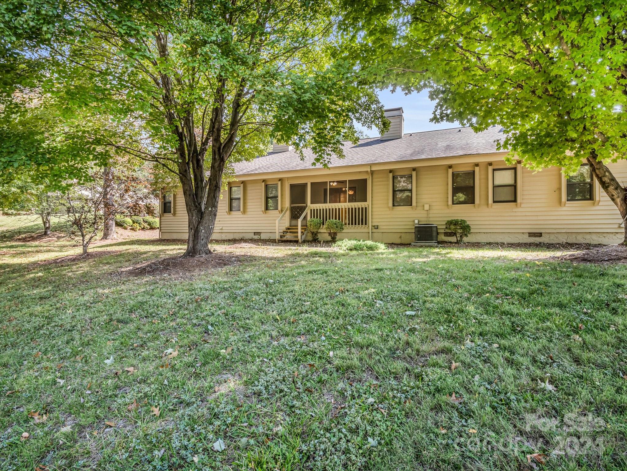 Single-story beige house with a small porch, framed by mature trees and a broad lawn.