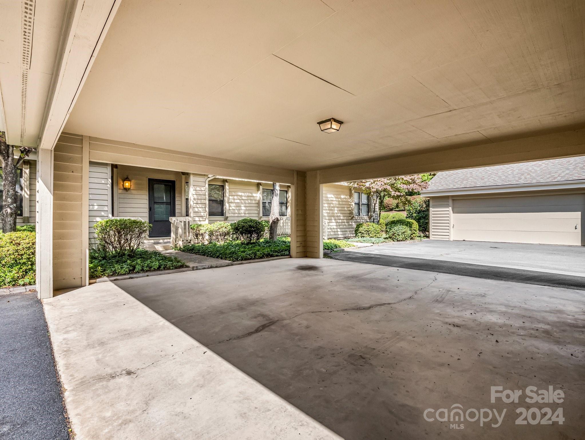 Covered carport beside the home’s entrance, with landscaped beds and a garage across the driveway.