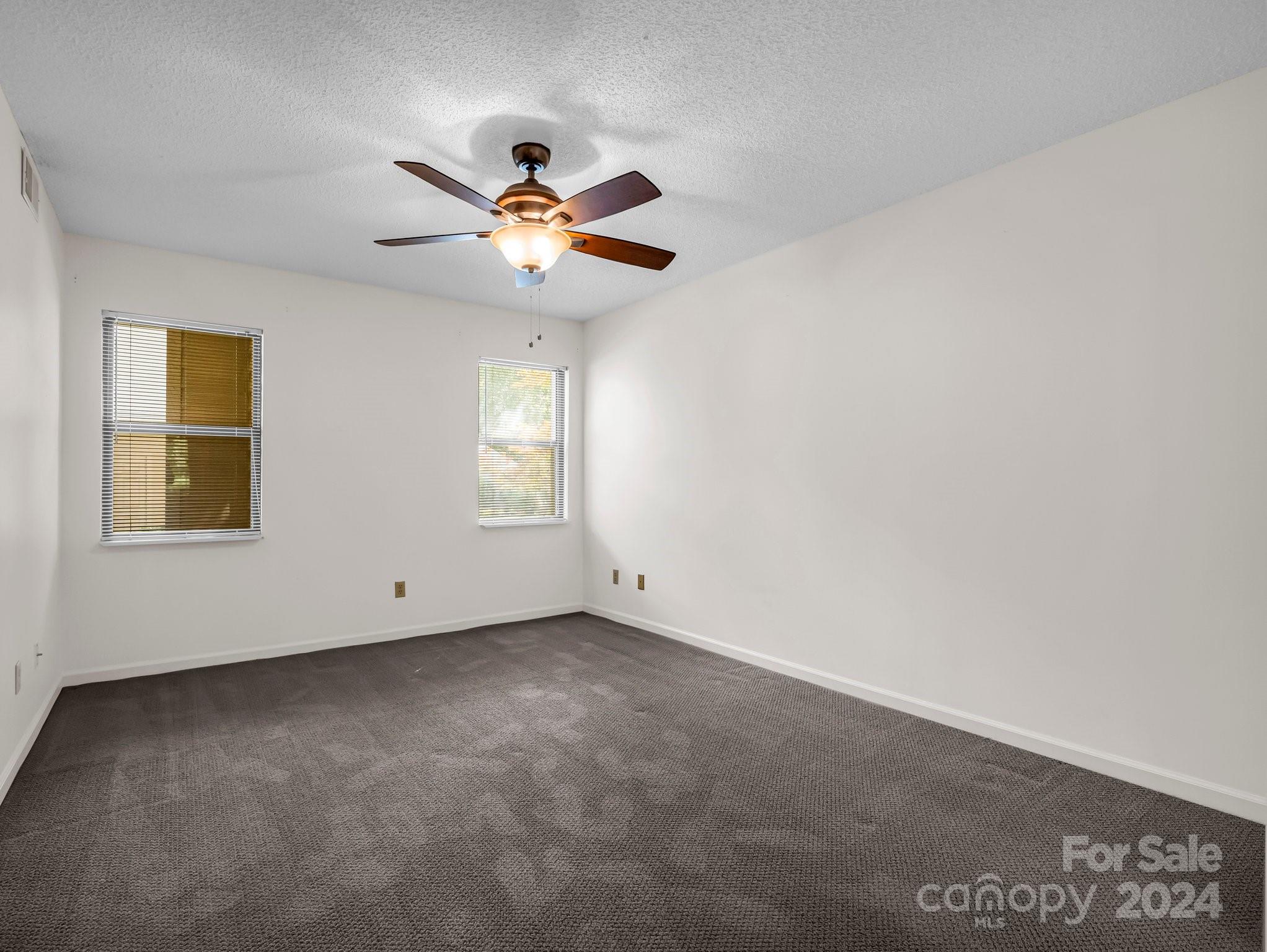 Empty carpeted bedroom with two windows and a lit ceiling fan.