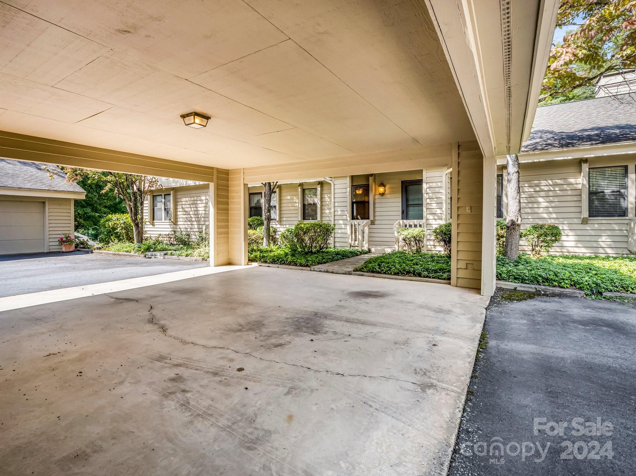 Covered carport and concrete parking area beside a light-sided home, with the landscaped front entrance beyond.