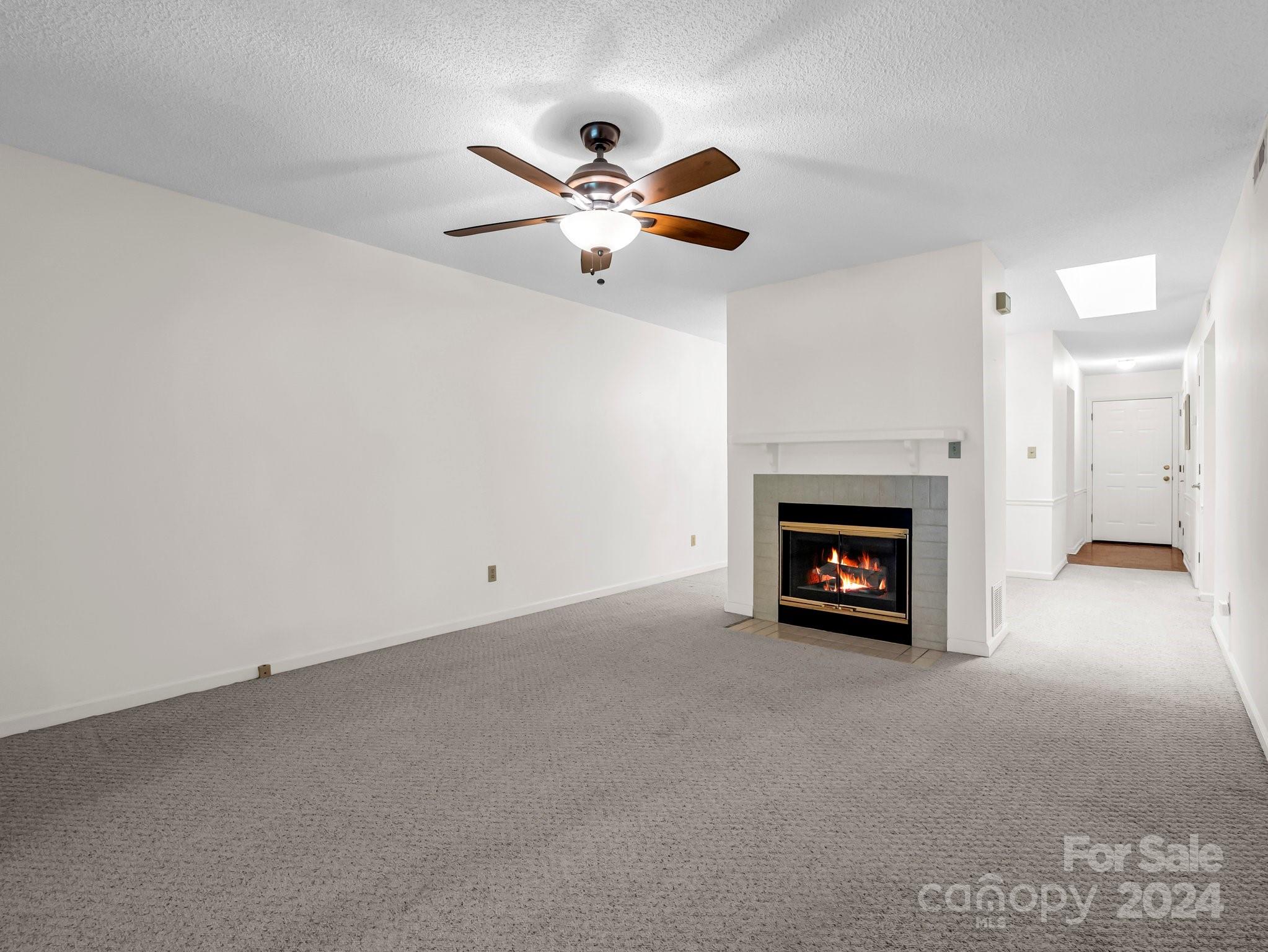Empty living room with a lit fireplace, gray carpet, and ceiling fan.