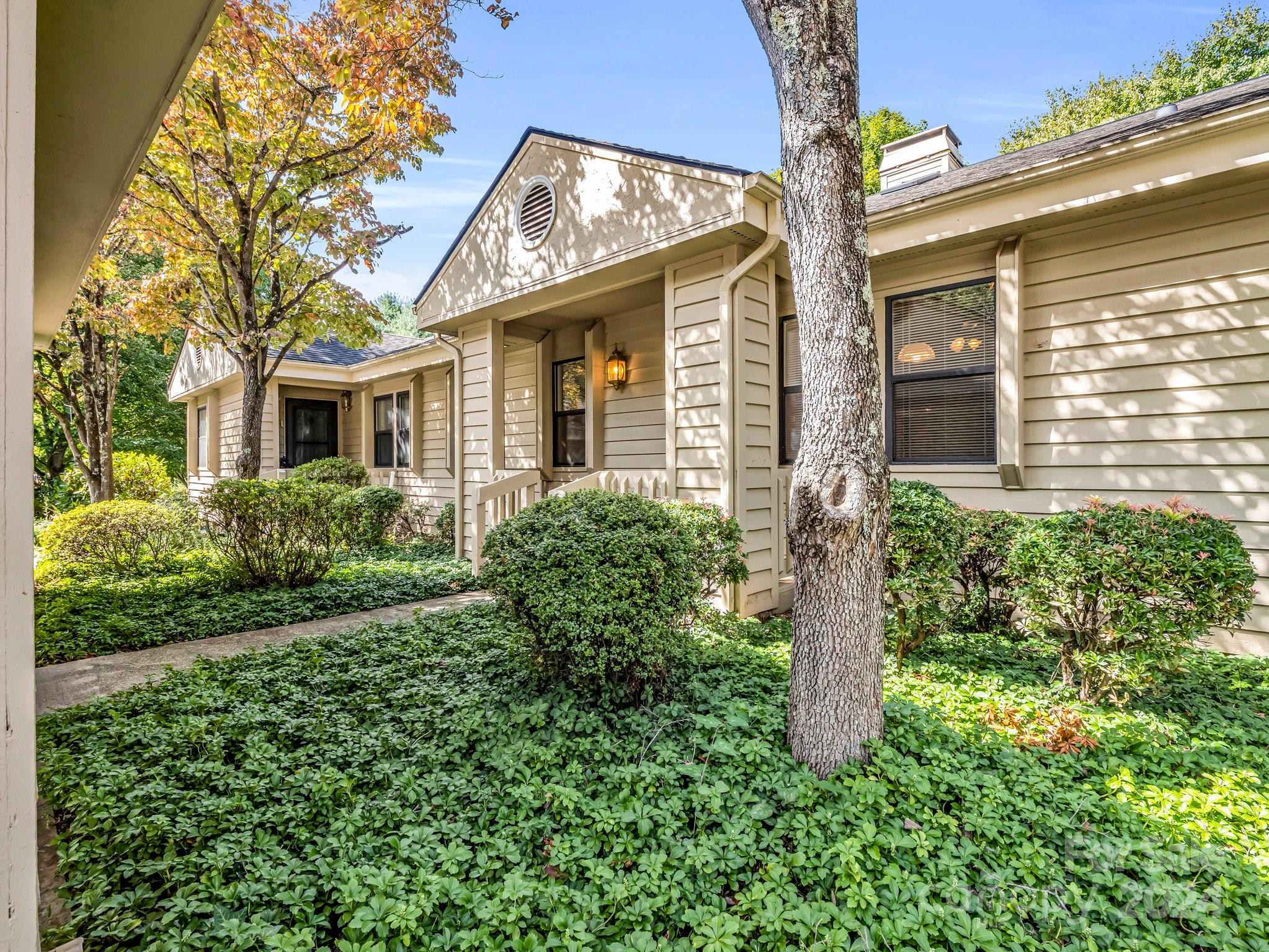 Beige-sided home with a covered entry, shaded by mature trees and surrounded by shrubs and groundcover.