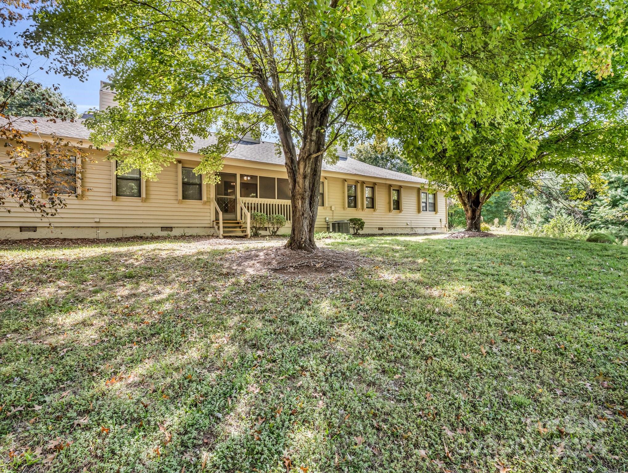 Single-story beige house with an entry porch, shaded by mature trees above a grassy lawn.
