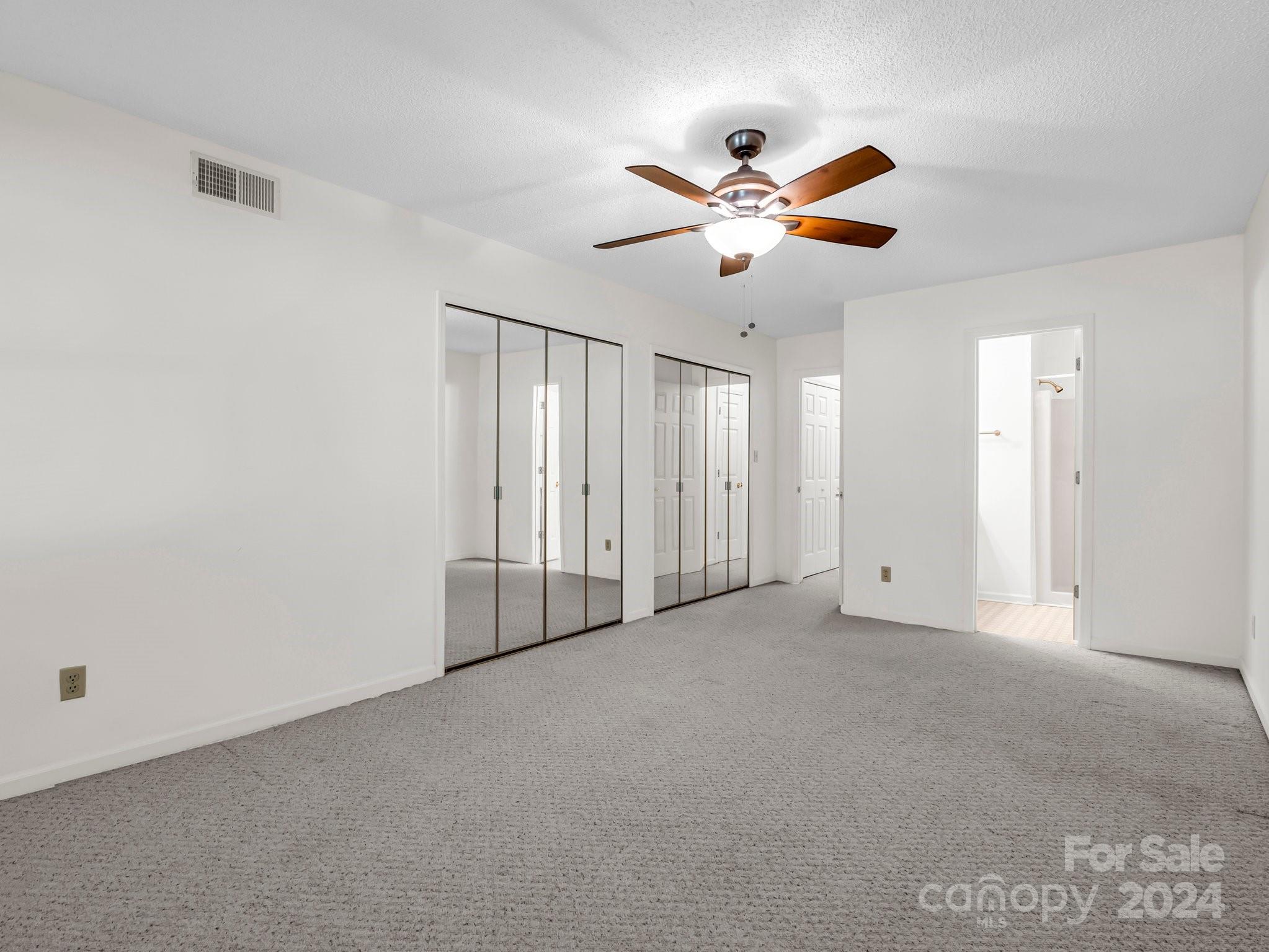 Empty bedroom with gray carpet, mirrored closet doors, and a wood-blade ceiling fan.