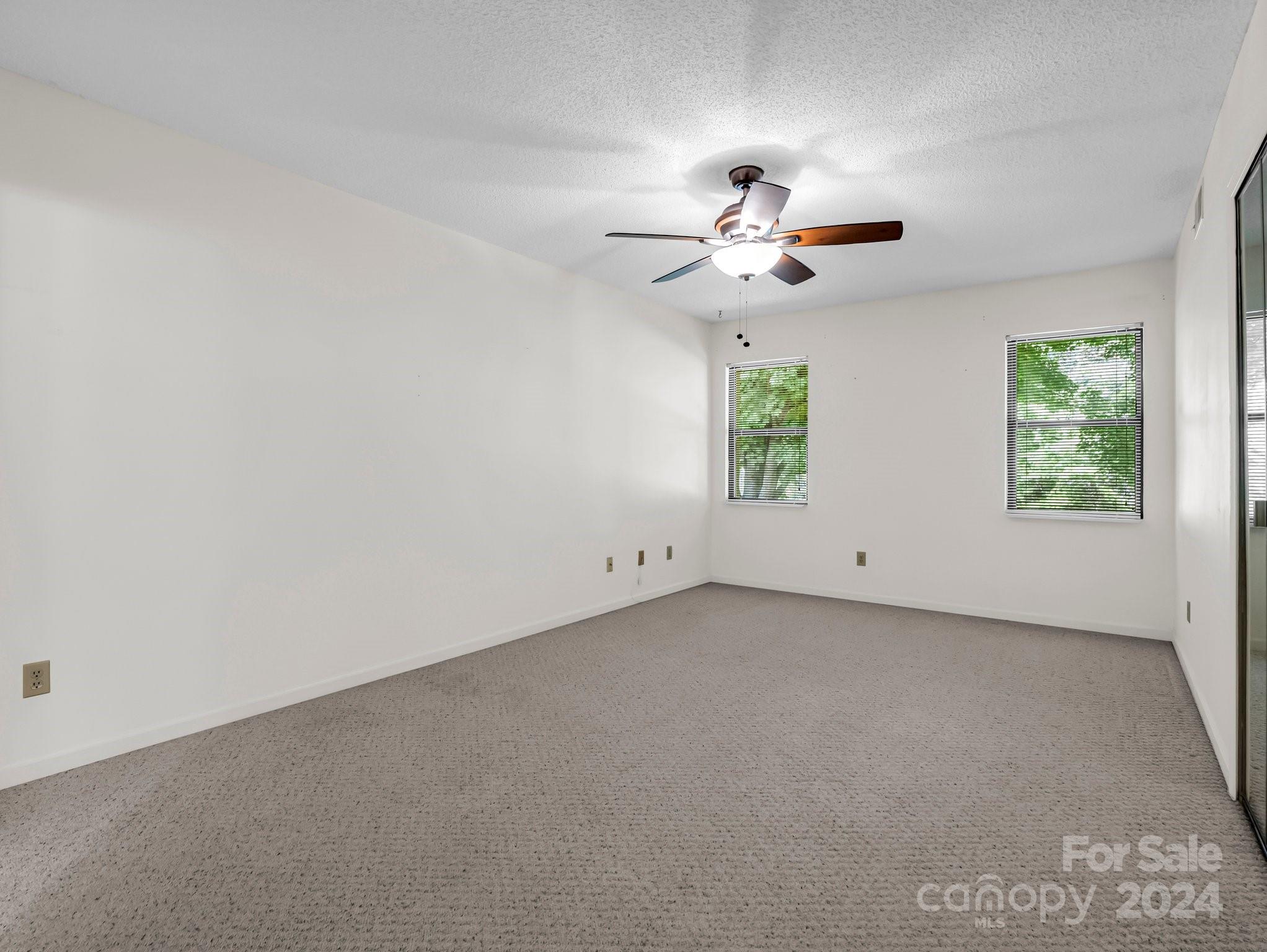 Empty carpeted room with a ceiling fan and two windows with blinds.