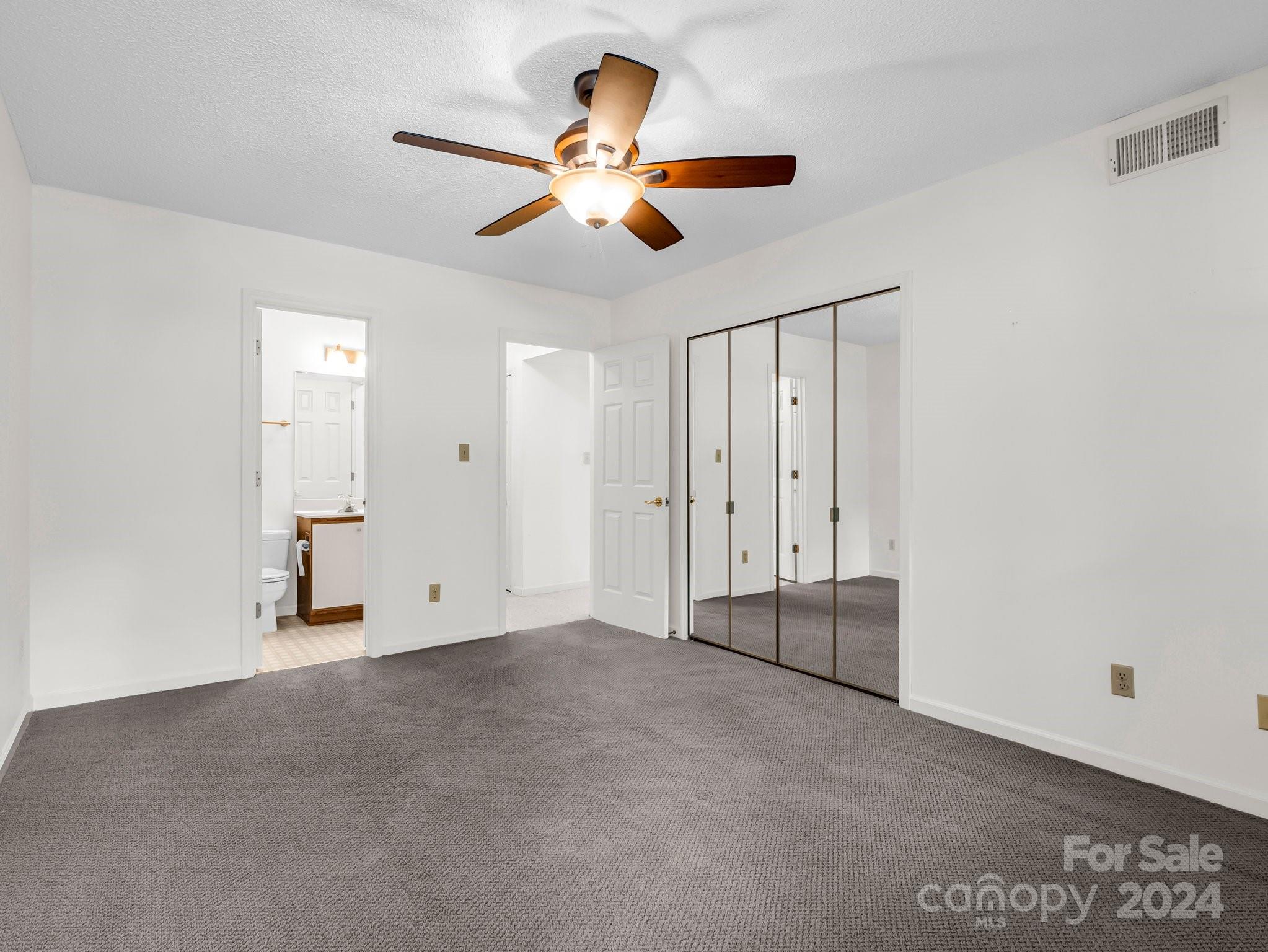 Empty bedroom with gray carpet, mirrored closet doors, a ceiling fan, and an adjoining bathroom.