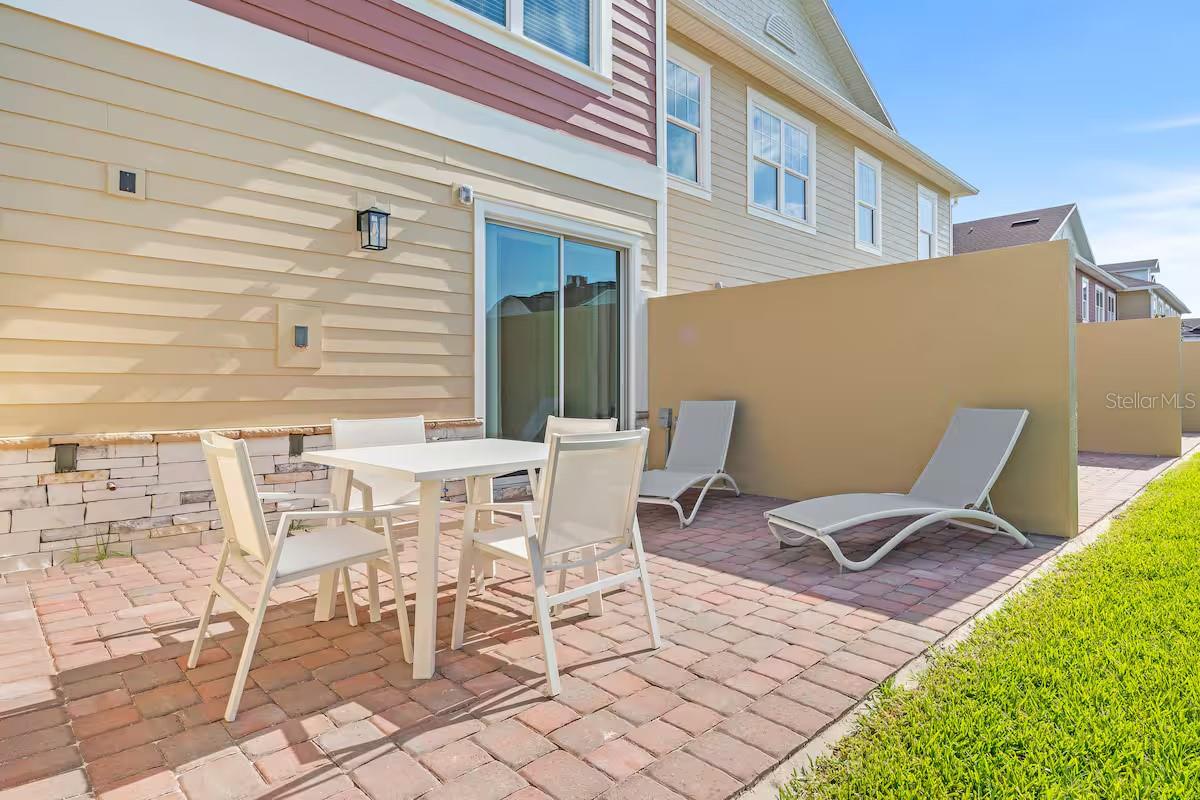 Brick-paver patio with white dining furniture, lounge chairs, and a tan privacy screen beside the home.