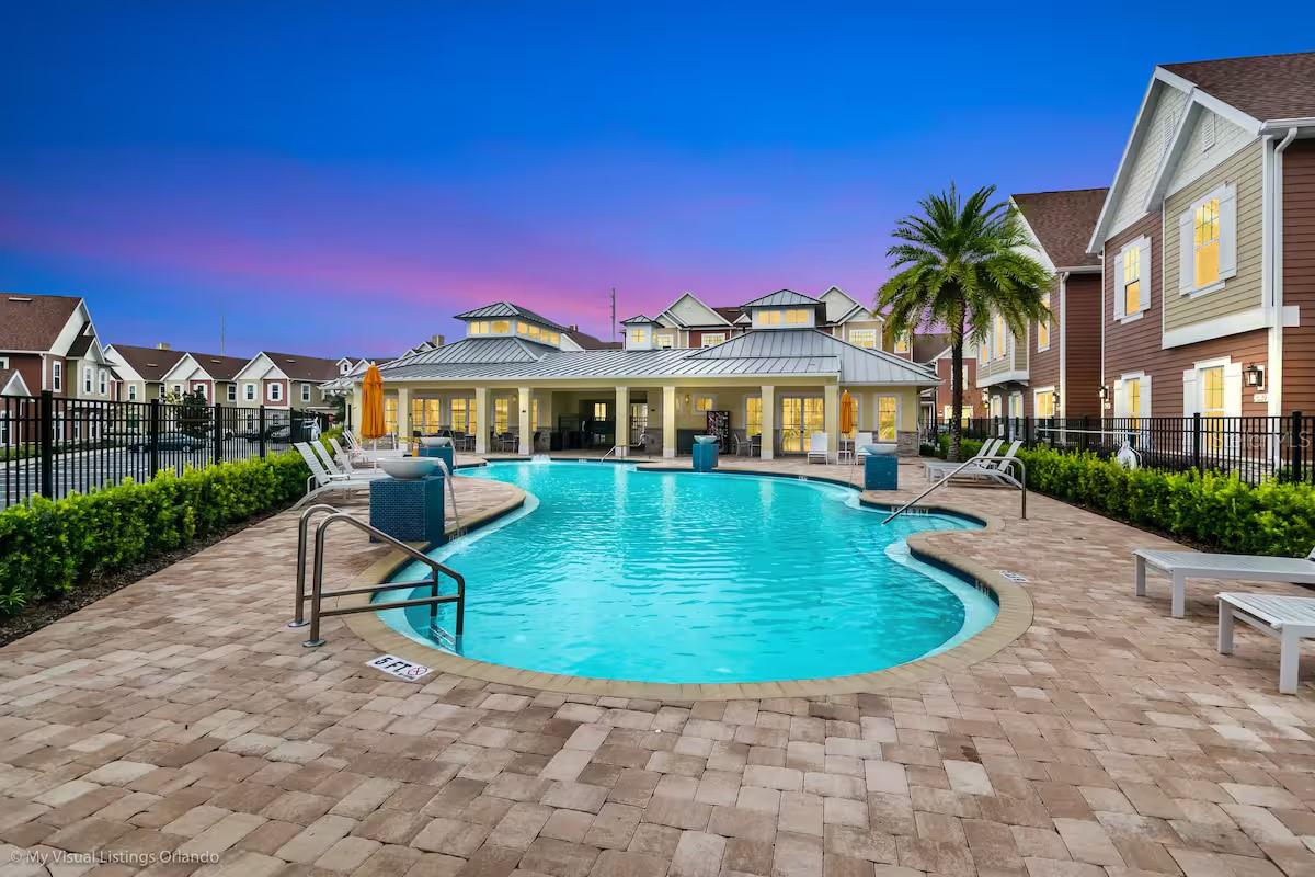 Community swimming pool with a curved shape, paved deck, clubhouse, and palm tree at dusk.