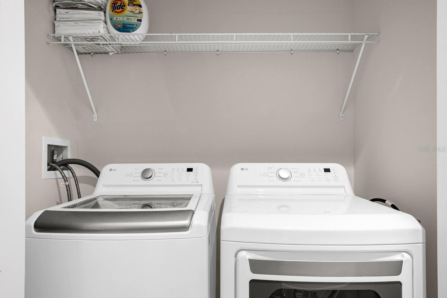 Laundry room with white top-loading washer and dryer beneath a wire shelf holding detergent and linens.