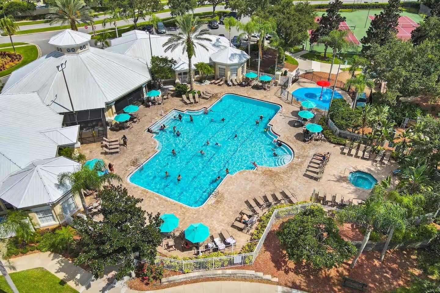 Aerial view of a community swimming pool with turquoise umbrellas, a clubhouse, and tennis courts.