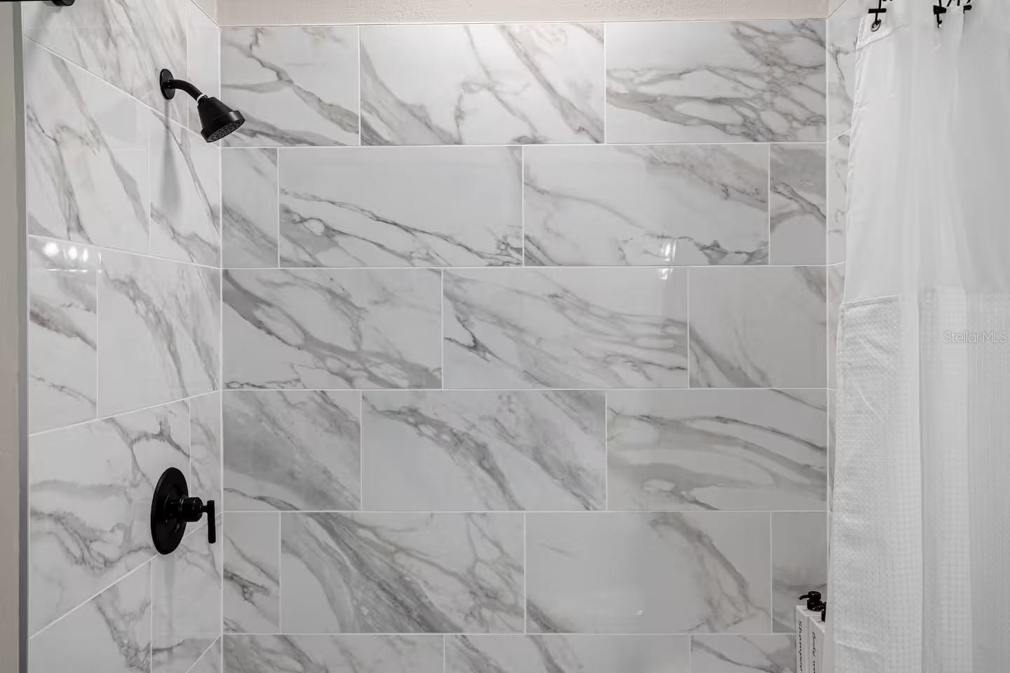 Shower with veined white tile walls, black fixtures, and a white curtain.
