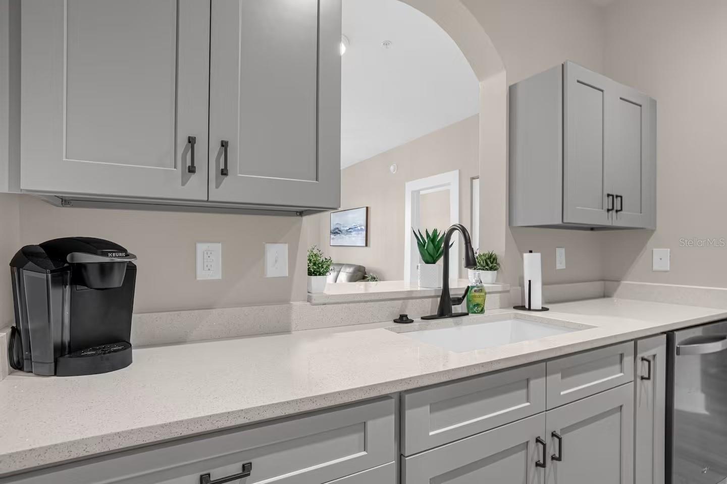 Kitchen with gray cabinets, white counters, black faucet, and coffee maker beside a pass-through to the living area.
