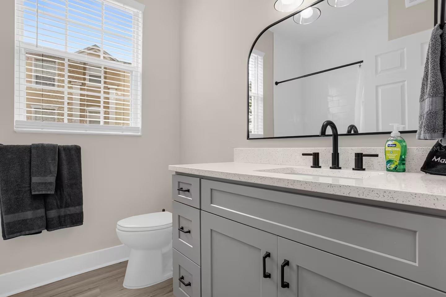 Bathroom with gray vanity and black faucet beside a toilet and bright window.
