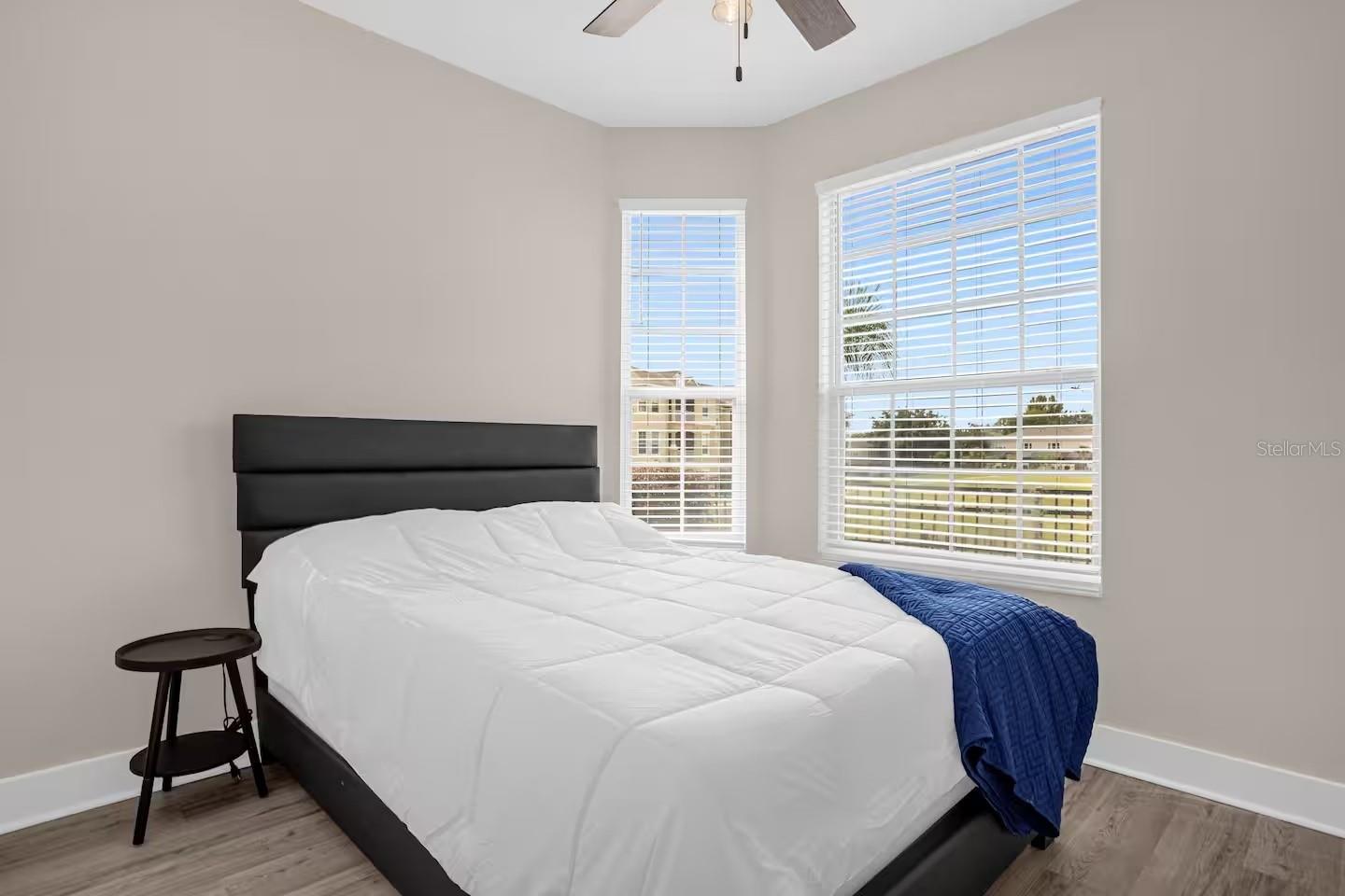 Bedroom with a large bed, blue throw, wood-look flooring, and two bright windows.
