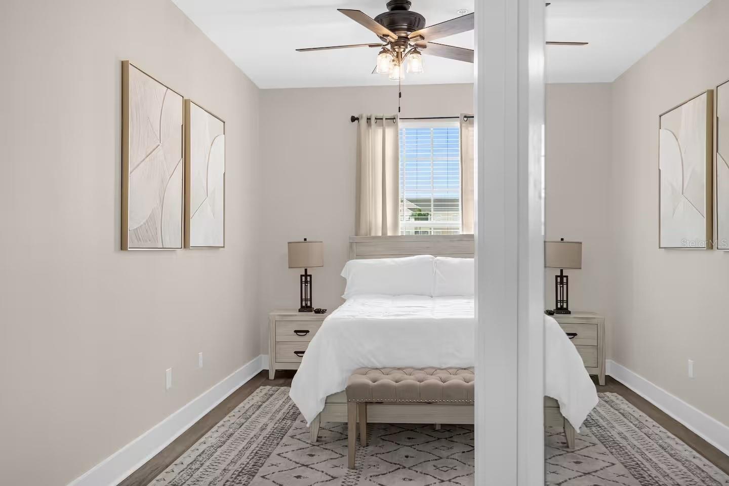 Staged bedroom with a white bed, ceiling fan, matching nightstands, and abstract wall art.
