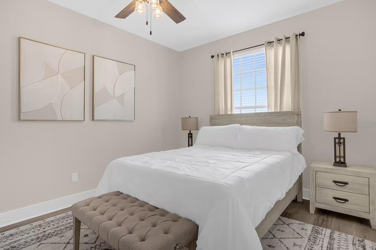 Staged bedroom with a white bed, tufted bench, light wood furniture, and a bright curtained window.