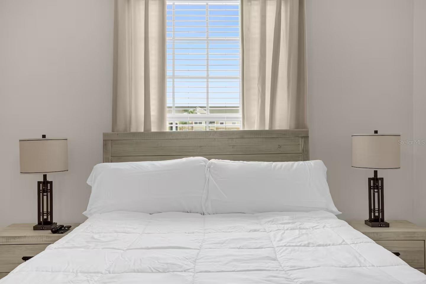 Bedroom with a white bed, light wood furniture, and a tall window behind the headboard.