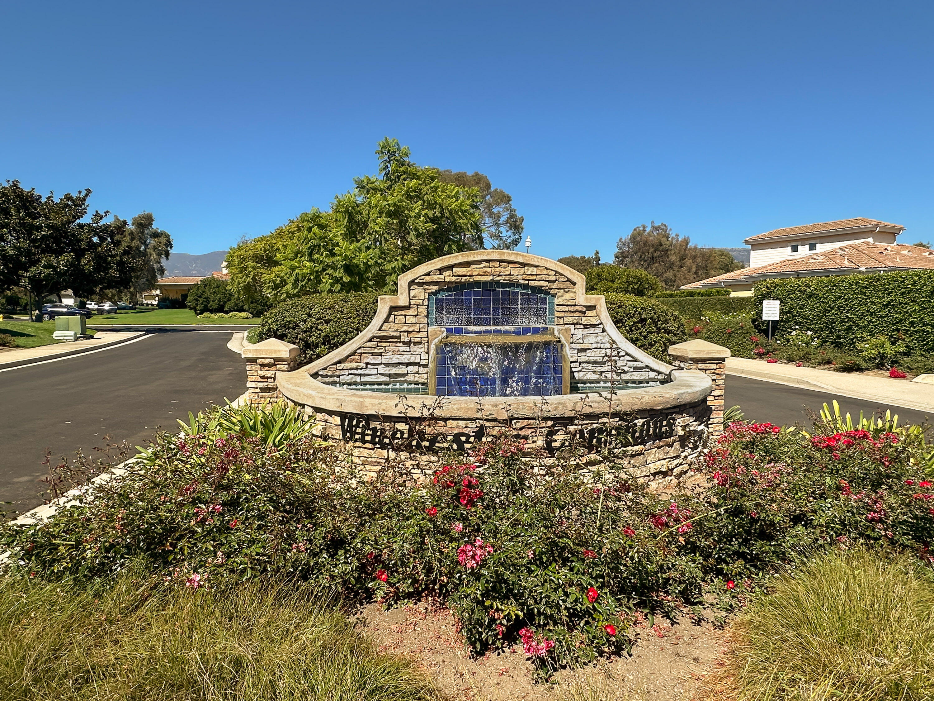 Stone community fountain with blue tile, surrounded by flowering shrubs and landscaped grounds.