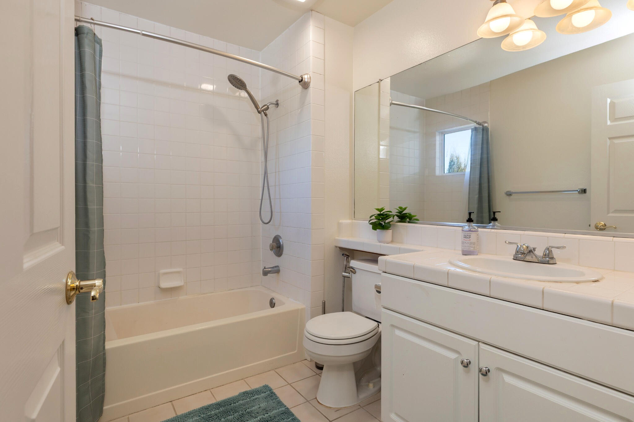 Bathroom with a tiled tub and shower, white vanity, large mirror, and blue-green bath mat.