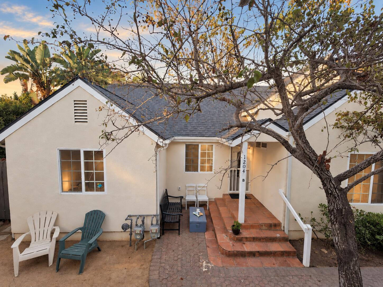 Stucco home with a tiled entry patio, outdoor chairs, and a large tree across the foreground.