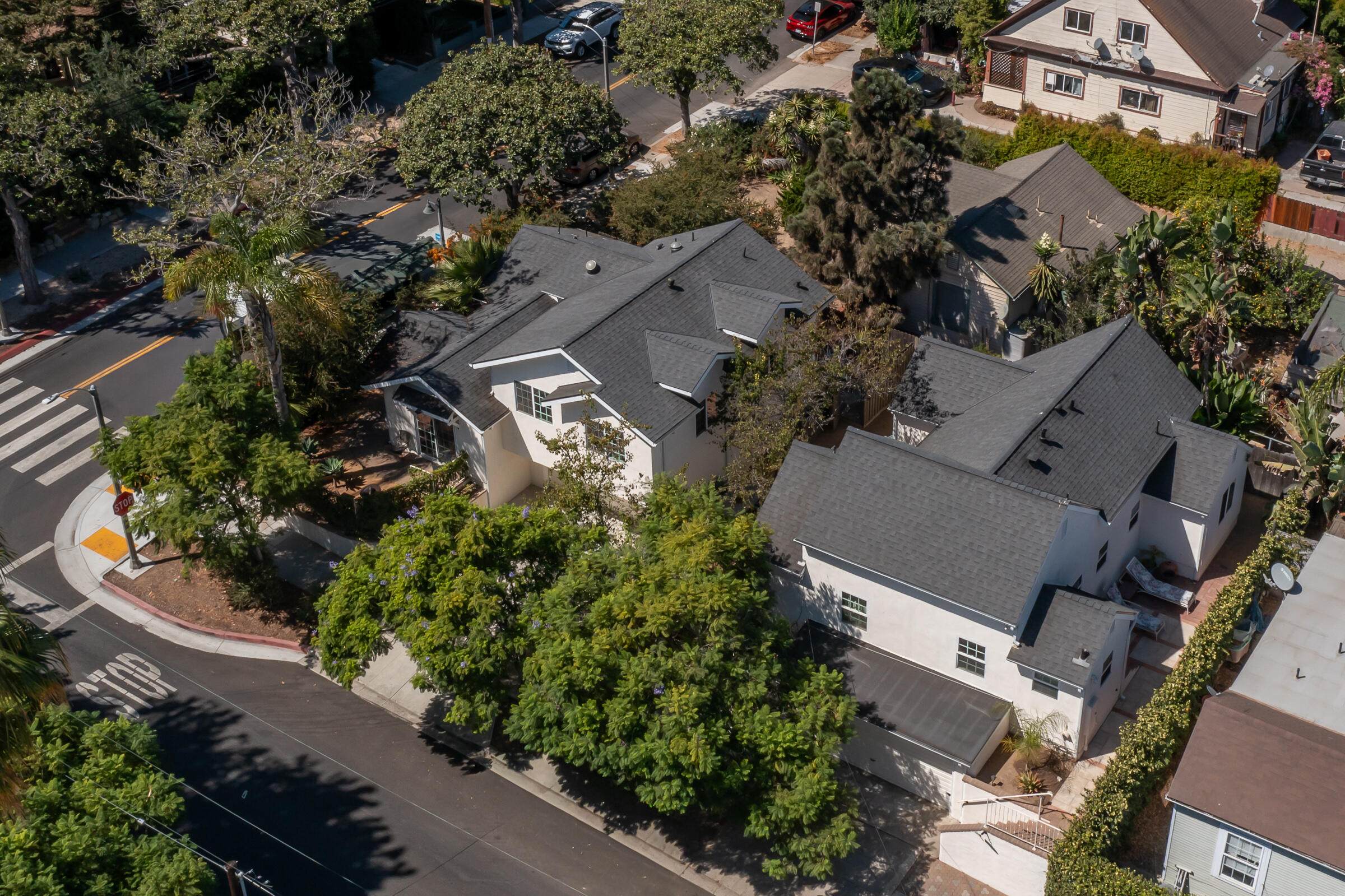 White home with dark gray roofs on a leafy corner lot, viewed from above.