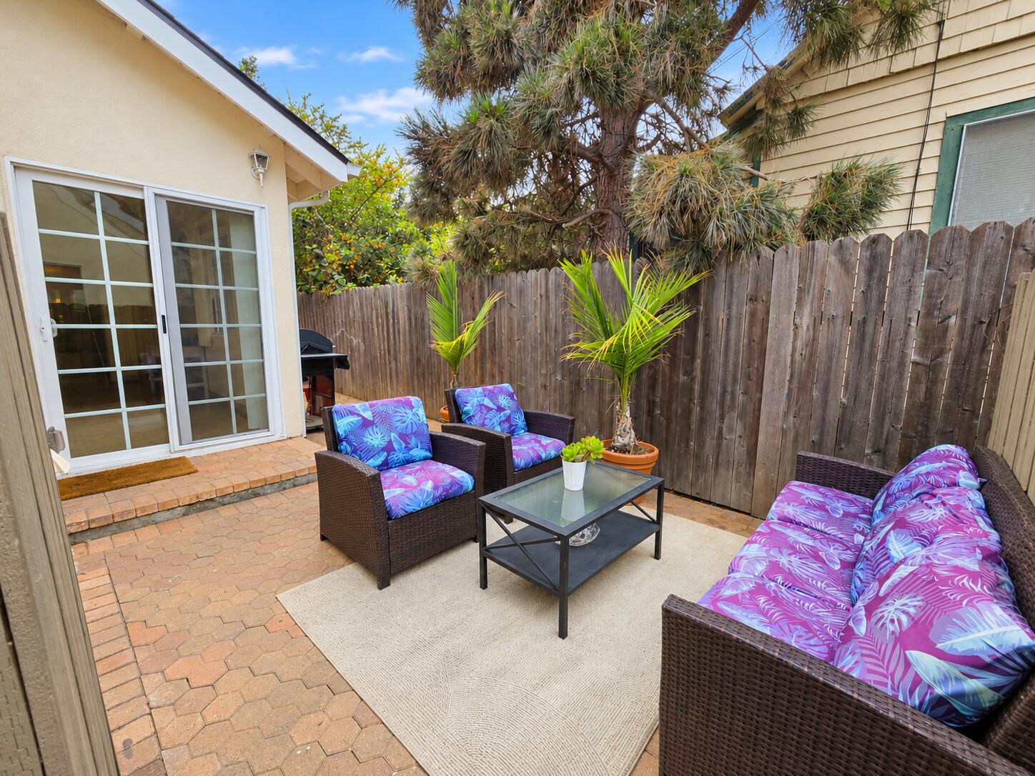 Private patio with wicker seating, purple floral cushions, potted palms, and a wooden privacy fence.