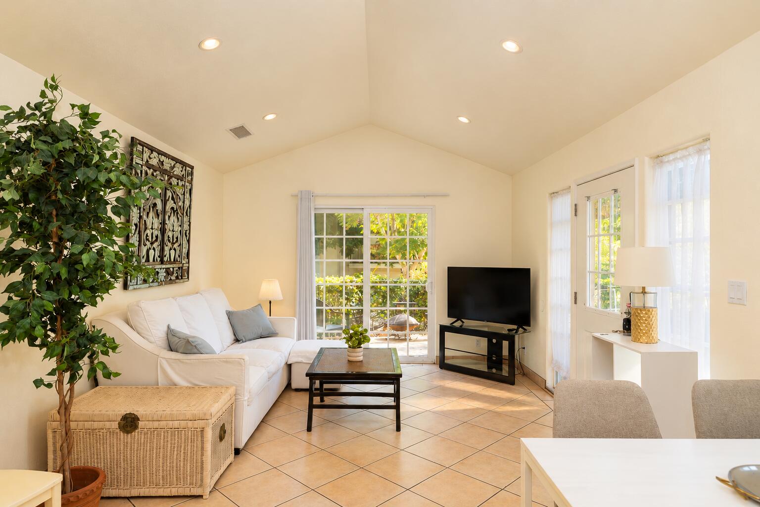 Living room with a white sectional, vaulted ceiling, tiled floor, and garden views through sliding glass doors.