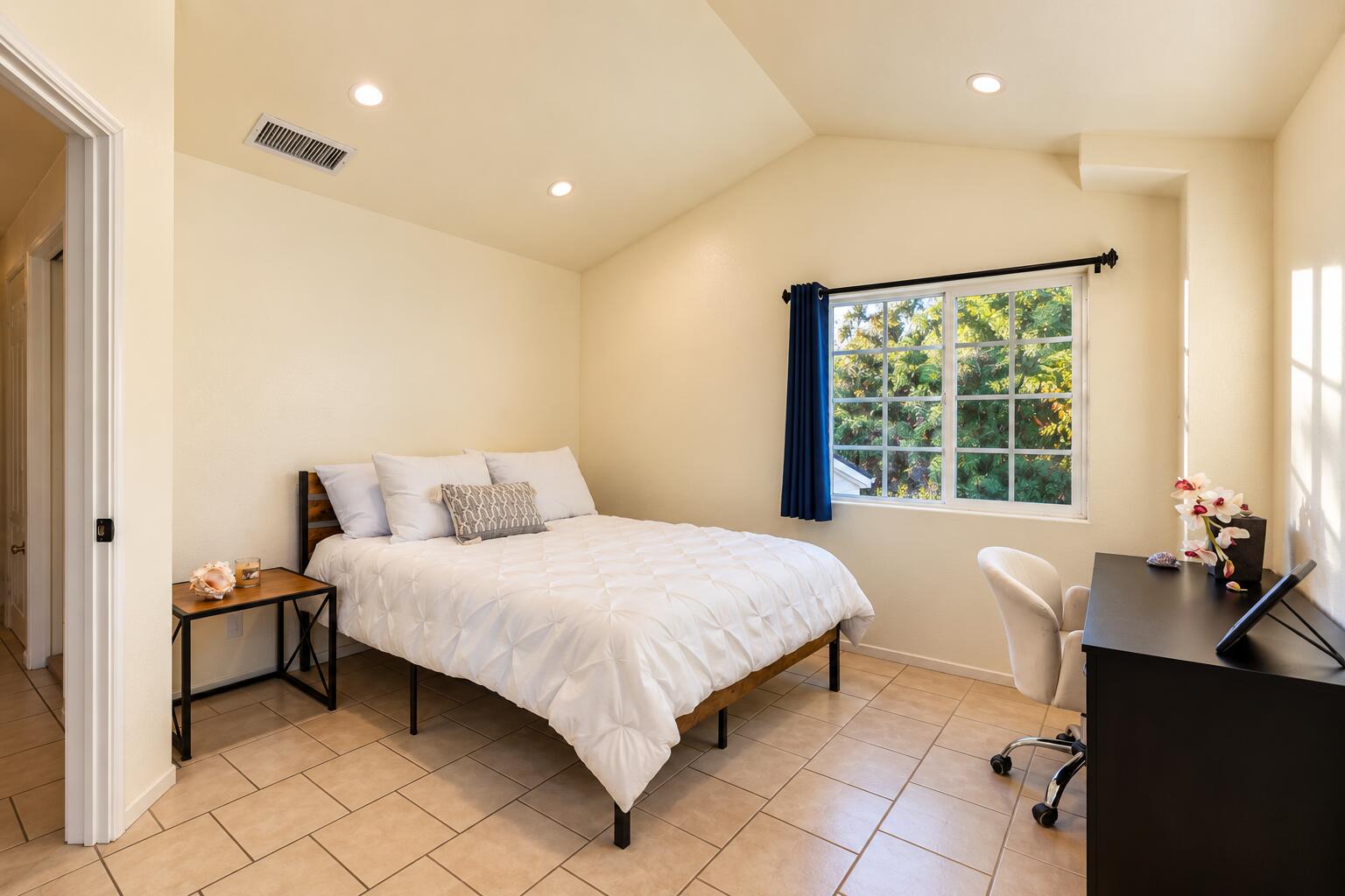 Bedroom with a white bed, blue curtains, and a desk beside a window overlooking greenery.