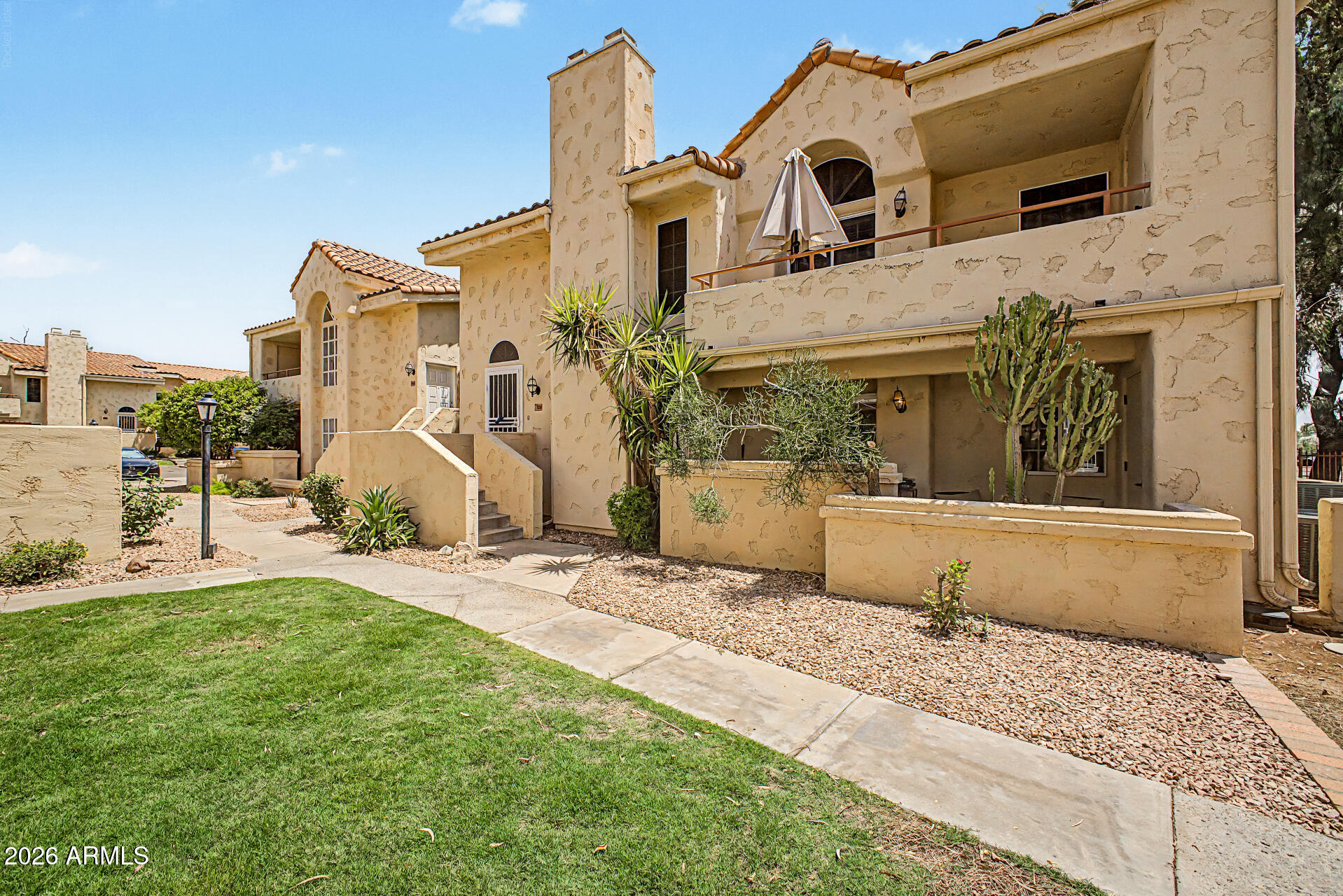 Tan stucco condominium building with tiled roofs, balconies, and desert landscaping.