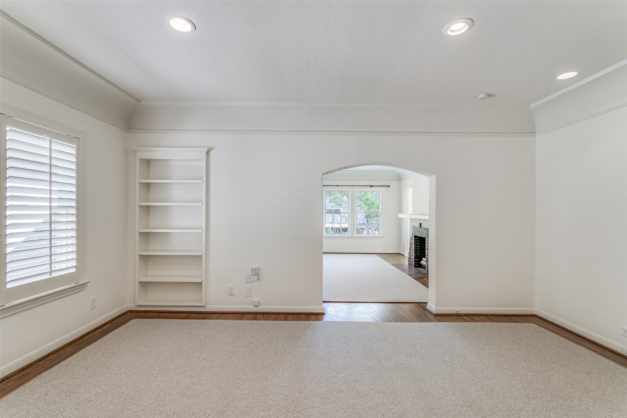 Empty carpeted room with built-in shelving and shutters, opening to a room with a brick fireplace.