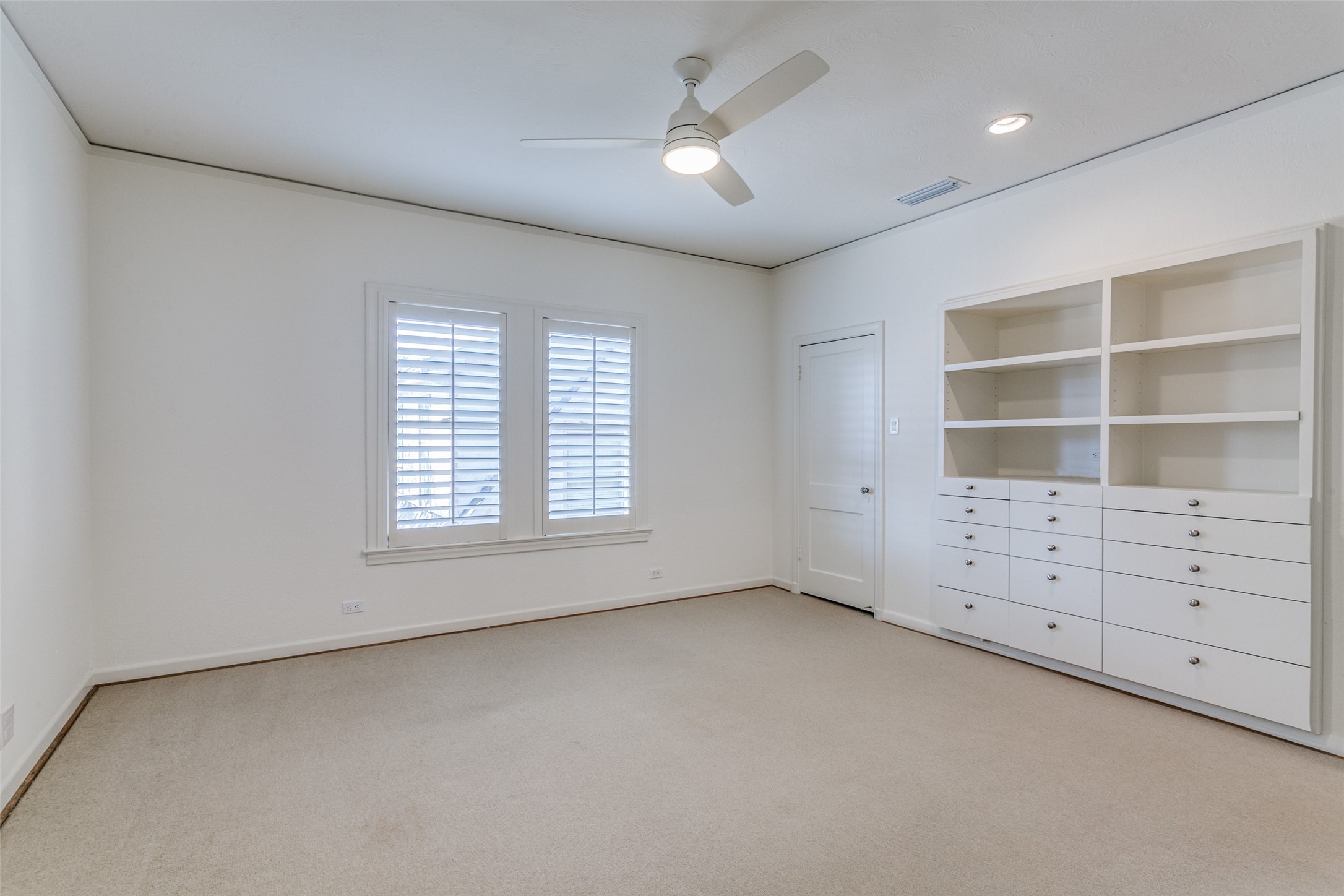 Empty bedroom with shuttered windows, beige carpet, and built-in white shelves and drawers.