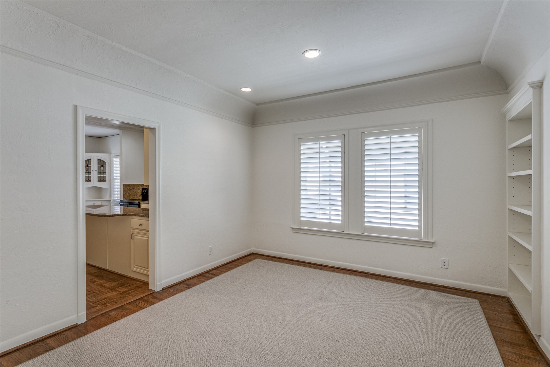 Empty room with plantation shutters, built-in shelves, and a large rug over hardwood floors.