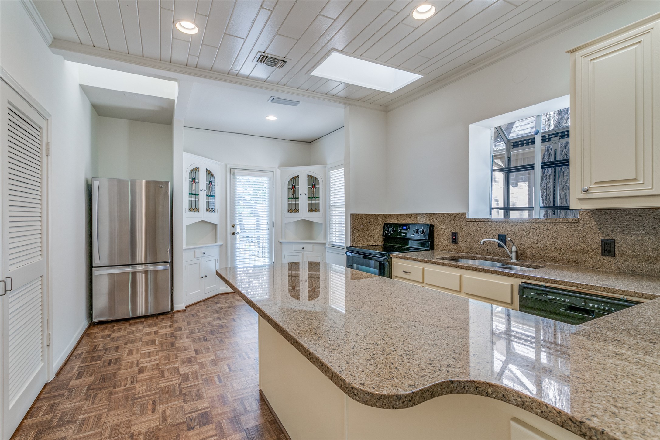 Kitchen with a curved granite island, stainless refrigerator, stained-glass cabinet doors, and parquet-pattern flooring.