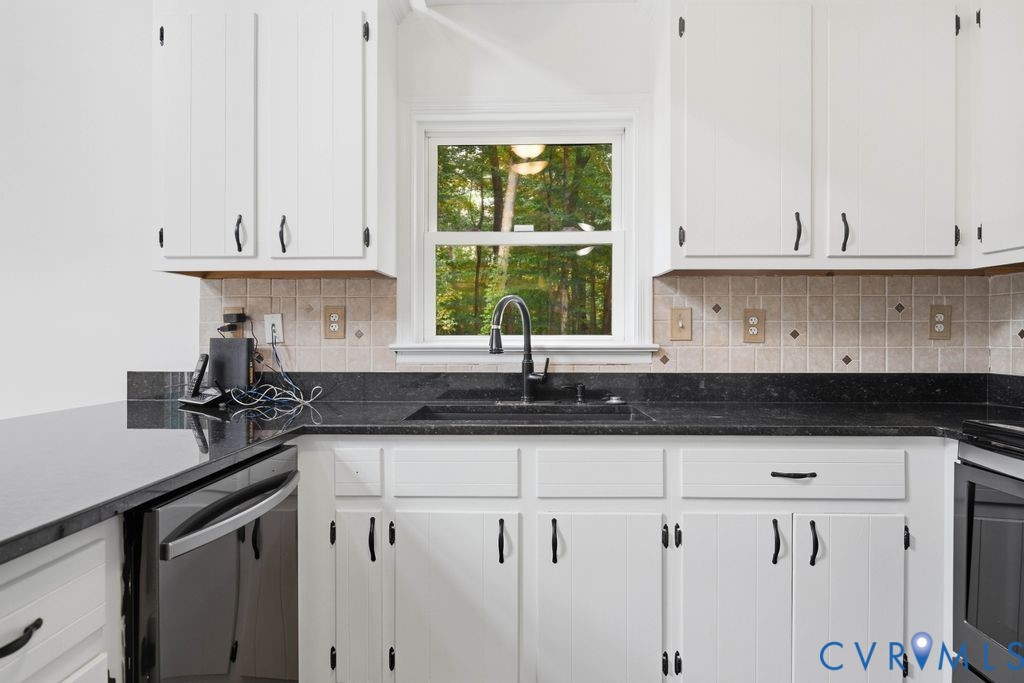 Kitchen with white cabinets, black countertops, and a sink beneath a window overlooking trees.