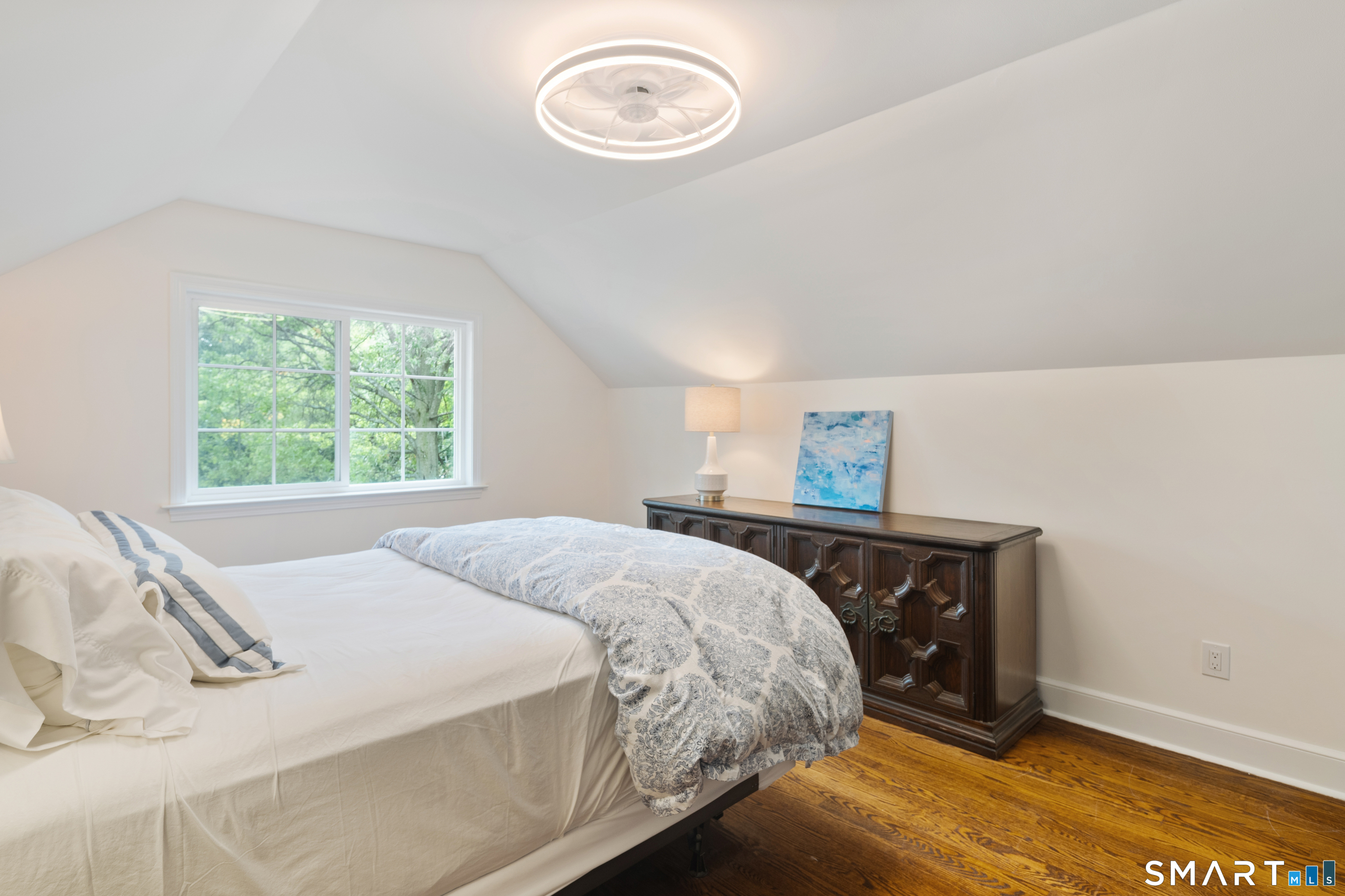 Furnished bedroom with a sloped ceiling, patterned bedding, hardwood floors, and a window overlooking trees.